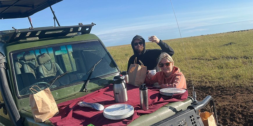 A woman and a man enjoying breakfast by a jeep in Tanzania